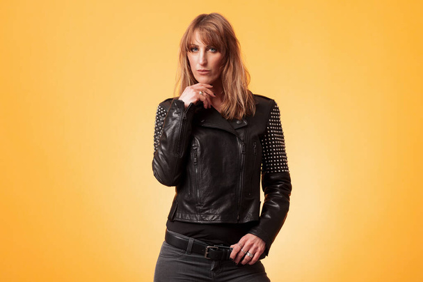 Portrait of an attractive blond woman wearing a studded leather jacket against a yellow background - Photo, image