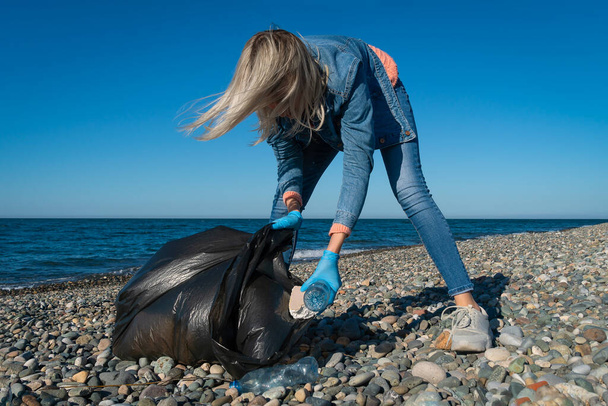 Γυναίκα συλλέγει πλαστικό, αφρό, καουτσούκ σε μια μαύρη σακούλα σκουπιδιών σε μια παραλία με βότσαλα στο φόντο της θάλασσας σε μια ηλιόλουστη μέρα. Καθαρισμός της παράκτιας ζώνης. Έννοια της οικολογίας, εθελοντισμός - Φωτογραφία, εικόνα