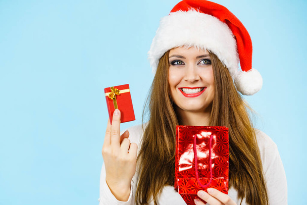 Christmas gifts. Happy woman wearing santa claus hat holding present red bag and small gift box with jewelery, on blue - 写真・画像