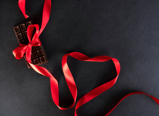 Natural organic bitter chocolate bar with red satin ribbon in a heart shape on black background. Happy Valentines day concept. - Фото, изображение
