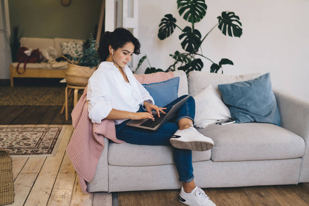 Side view young concentrated brunette in casual outfit and earphones text messaging on netbook while sitting with crossed legs on comfortable couch - Foto, imagen