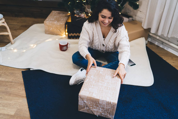From above of happy female in sweater opening box with gift while sitting on carpet under Christmas tree with garland - Zdjęcie, obraz