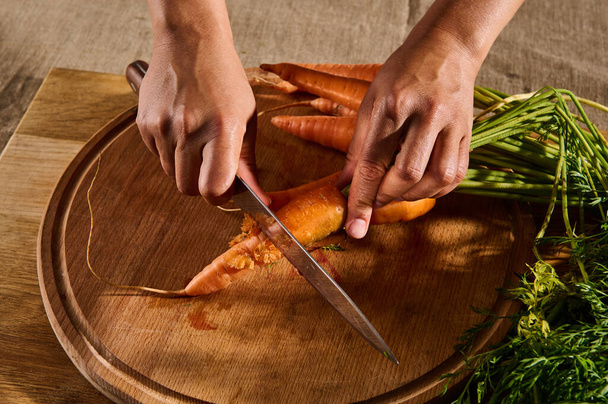 Bovenaanzicht van vrouwelijke handen snijden, hakken, snijden verse rauwe baby wortel op een houten plank. Close-up van de hand met mes snijden verse groenten. - Foto, afbeelding