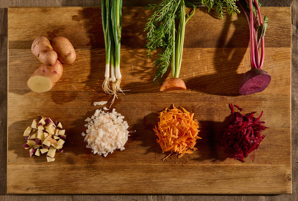 Overhead view of seasonal fresh raw vegetables on a wooden board, cut in half and the other half shredded on a grater. Beets, carrots, green onions with leaves and potatoes. Flat lay. Food background - Foto, imagen