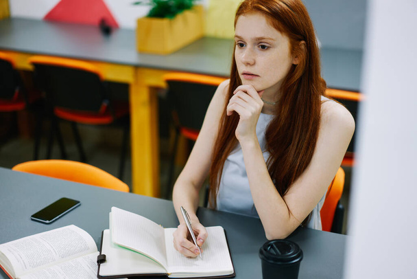 Koncentrált fiatal nő ül asztalnál a modern munkahelyen, és írni gondolatok notebook, miközben néz félre, és visel alkalmi ruhák, könyv és telefon az asztalnál - Fotó, kép