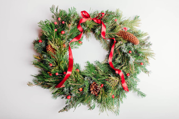 Christmas and New Year wreath made of fir, pine spruce evergreen branches, rose hips, cones decorated with red ribbon bow on white background. - Zdjęcie, obraz