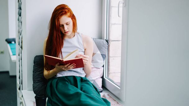 Concentrated young woman wearing casual clothes while sitting comfortably on windowsill and reading interesting novel in light room in daytime - Photo, Image