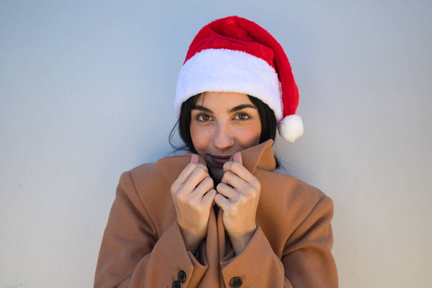 Jonge, mooie, donkerharige vrouw met een kerstmuts en een bruine jas om zichzelf te beschermen tegen de kou. De vrouw maakt verschillende houdingen. Concept vrolijk kerstfeest en gelukkig nieuwjaar. - Foto, afbeelding