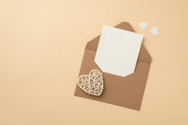 Photo de vue du dessus de l'enveloppe en papier artisanal ouvert avec feuille de papier coeurs blancs et coeur en rotin sur fond beige isolé avec copyspace - Photo, image