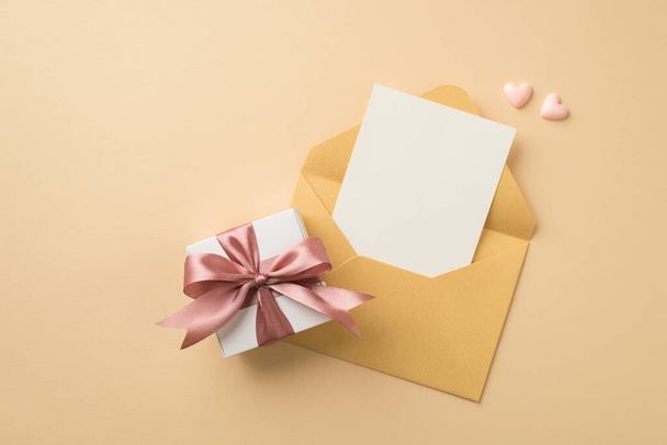 Фото открытого пастельного желтого конверта с карточкой маленьких розовых сердец и белой коробочкой подарков с светло-розовым бантом на изолированном бежевом фоне с копирайтом - Фото, изображение