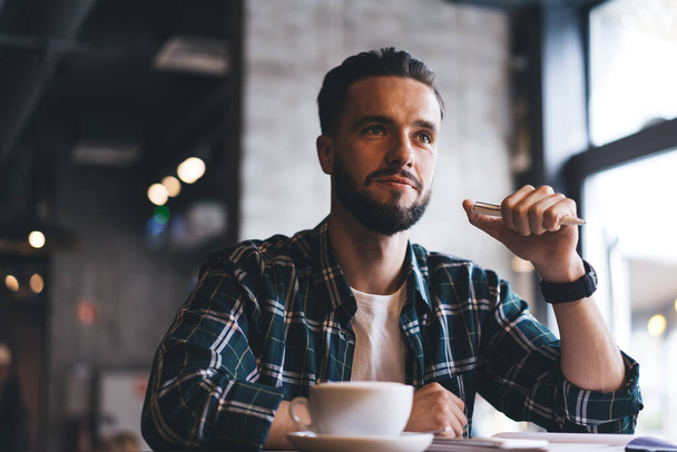 Contemplative male poet with pen in hand thinking about creative idea during free time in cafe interior, pensive hipster guy with caffeine cappuccino sitting at cafeteria table and feeling pondering - Foto, Imagen