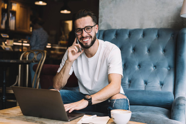 Portrait of cheerful male programmer in optical spectacles smiling at camera during positive cellphone conversation for discussing web networking, happy Caucaisan blogger using 4g for calling and talk - Φωτογραφία, εικόνα