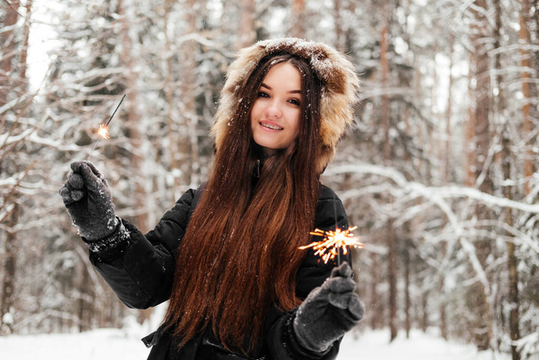 Egy fekete kabátos nő kezében csillogóval és mosolyogva nézi a kamerát. 20 éves lány a téli erdőben szabadban - Fotó, kép
