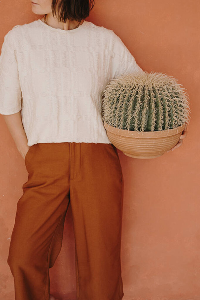 Mujer permanecer contra la pared de jengibre melocotón y mantener cactus planta en casa - Foto, imagen