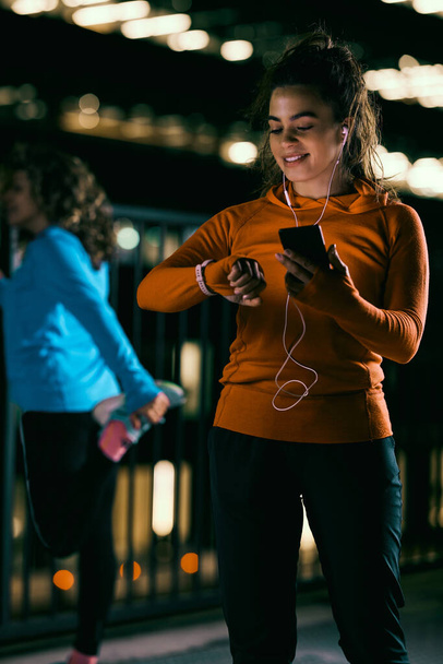 A fit sportswoman is standing on the street with headphones and a phone and looking at her smartwatch. She is checking on her heart rate on the app. - Foto, immagini