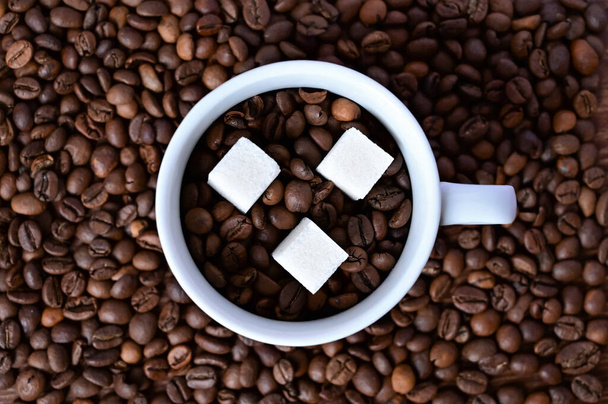white cup with coffee beans and three pieces of sugar on a coffee background. Close-up. Flat layout with top view - Foto, Imagem