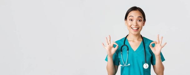 Gesundheitshelfer, Virusprävention, Kampagnenkonzept für Quarantäne. Aufgeregt und erfreut asiatische Ärztin, Krankenschwester in Peelings mit Stethoskop, zeigt okay Geste, weißer Hintergrund - Foto, Bild