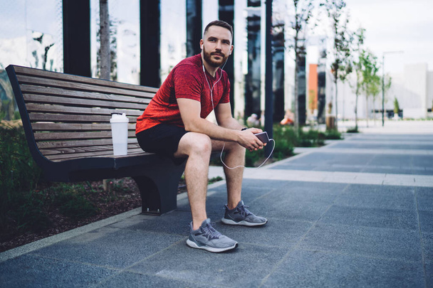 Full body of confident male athlete in earphones sitting on bench and looking away while chilling after morning training in city - Фото, изображение
