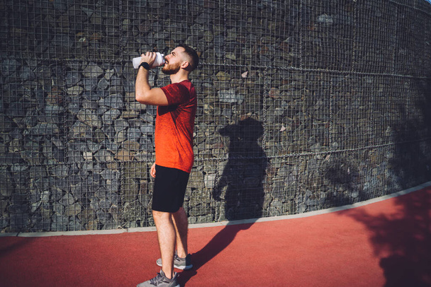 Boční pohled na seriózního vousatého muže stojící na sportovním hřišti a popíjejícího z plastové láhve při odpočinku po cvičení fitness lekce - Fotografie, Obrázek