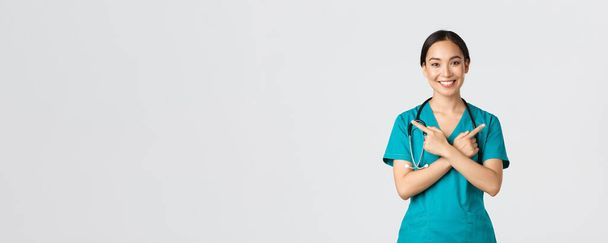 Covid-19, gezondheidswerkers, pandemisch concept. Lachende mooie aziatische verpleegster, stagiaire of arts in scrubs wijzen zijwaarts en kijken naar de camera, tonen varianten, twee keuzes, witte achtergrond - Foto, afbeelding