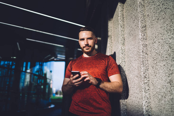 Young bearded male wearing red shirt surfing mobile phone and looking at camera while leaning on wall on sunny summer day - Photo, Image
