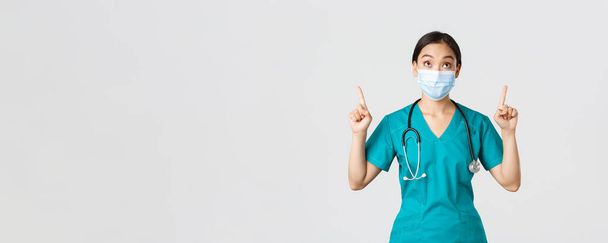 Ковид-19, коронавирусная болезнь, концепция медицинских работников. Заинтригованная и взволнованная азиатка врач, медсестра в медицинской маске и перчатках, смотрящая и указывая пальцами вверх, белый фон - Фото, изображение