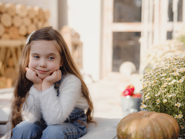 Happy little girl sitting on porch of house. home fall decoration for Thanksgiving. Smilling Child in autumn garden with yellow pumpkins - Photo, Image