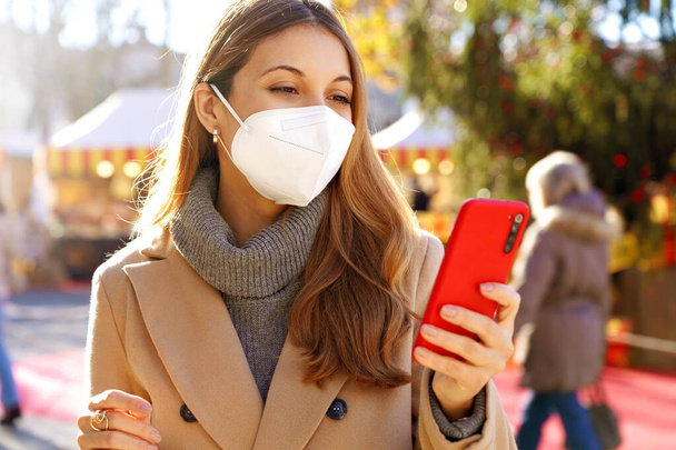Beautiful young woman wearing medical face mask using smartphone on Christmas time outdoors - Foto, immagini