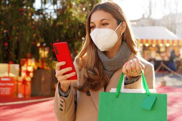 KN95 FFP2 maskeli şık bir kadın elinde alışveriş torbalarıyla Noel pazarları arasında yürürken telefonunu izliyor. - Fotoğraf, Görsel