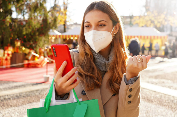 Mujer joven emocionada con máscara médica FFP2 KN95 viendo smartphone y sosteniendo bolsas de compras caminando en los mercados de Navidad - Foto, imagen