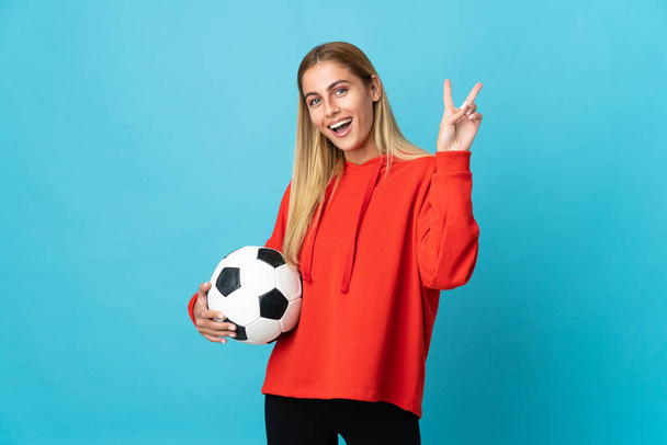 Fiatal labdarúgó nő elszigetelt kék háttér mosolyog és mutatja győzelem jele - Fotó, kép