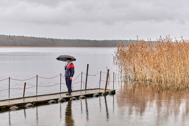 Ασιάτης άνθρωπος με τα πόδια κάτω από ομπρέλα σε ξύλινη προβλήτα το φθινόπωρο βροχερή γκρι ημέρα πάνω από το σκηνικό της λίμνης και του δάσους. - Φωτογραφία, εικόνα