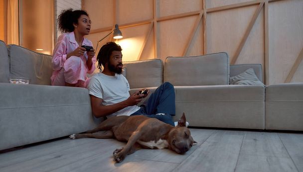 Чорна пара грає в відеогру вдома біля собаки
 - Фото, зображення