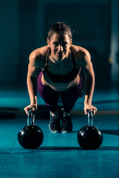 A young woman in a gym doing push-ups with the help of a ball-shaped weight known as a kettlebell. - Photo, Image