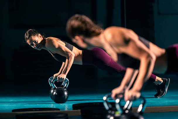 Uma jovem mulher em um ginásio fazendo flexões com a ajuda de um peso em forma de bola conhecido como kettlebell. - Foto, Imagem