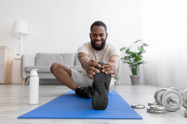 Glücklicher Millennial-Sportler mit schwarzem Bart macht Bewegung für Beine, dehnt sich auf dem Boden auf Matte mit Kurzhanteln - Foto, Bild