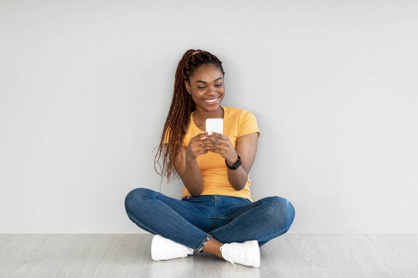 Schwarze Frau mit Mobiltelefon, Videochat im Web, Arbeit oder Studium online, auf dem Boden sitzend gegen graue Wand - Foto, Bild