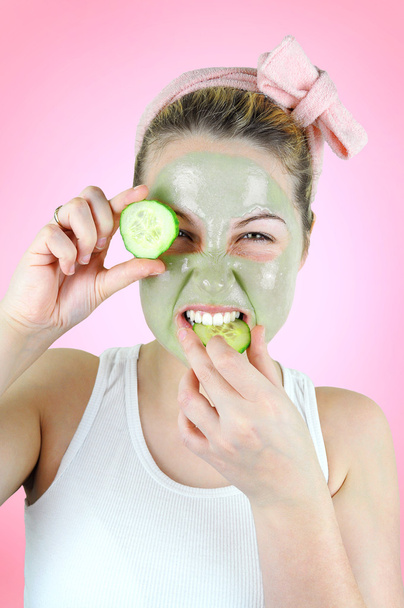 Beauty portrait of a funny woman wearing a pink headband, white top and a green facial mask is holding a slice of cucumber in front of her left eye and is about bite into another one. - Photo, Image