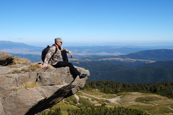 un hombre adulto se sienta con una mochila en el borde de un acantilado y mira soñadoramente a la distancia. - Foto, Imagen