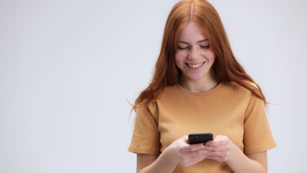Hübsche junge Frau mit Smartphone, während sie vor isoliertem weißen Hintergrund steht. Sie lächelt, hat Spaß an der Kommunikation in sozialen Netzwerken auf ihrem Handy - Filmmaterial, Video