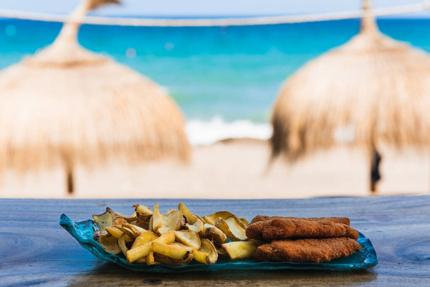 Blauer Teller mit Hühnchen und Chips vor dem Strand mit zwei Sonnenschirmen im Hintergrund - Foto, Bild