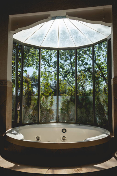 Increíble bañera de hidromasaje con hermosa ventana con vista al jardín con árboles, cielo azul y luz solar - Foto, imagen