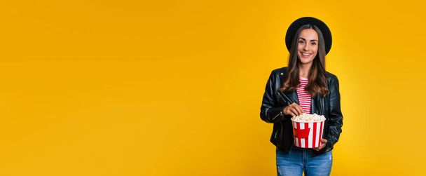 Banner photo of stylish young beautiful hipster woman in black jacket and hat with a bucket of popcorn while she is posing isolated on yellow background - Фото, изображение
