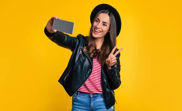 Close up portrait of happy trendy beautiful hipster woman in black jacket and hat while she is making selfie photo and isolated on yellow background - Foto, immagini