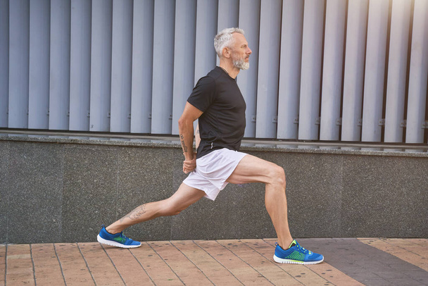 Homme d'âge mûr sportif confiant en vêtements de sport étirant ses jambes, se réchauffant et faisant de l'exercice à l'extérieur - Photo, image