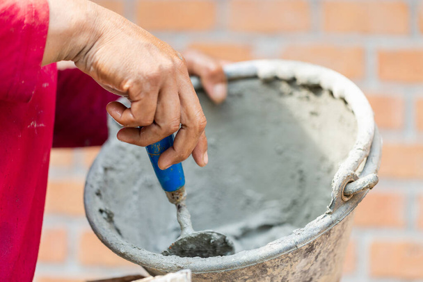 Закройте руку работника, держащего лопату и ведро со свежим цементом, строитель-каменщик, строящий стены из кирпича, раствора и шпатлевки - Фото, изображение