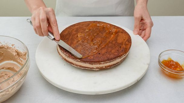 Assembling the chocolate cake. Woman hands spread the top of cake with apricot jam.  Step by step chocolate cake with chocolate cream recipe, close up baking process. - Fotó, kép