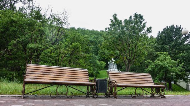 Na chodniku w parku znajdują się dwie drewniane ławki. Za bujną zieloną roślinnością znajduje się stara, szaro-biała kaplica. Pietropawłowsk-Kamczacki - Zdjęcie, obraz