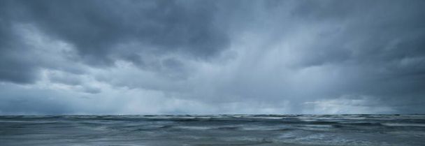 Темное штормовое небо над Балтийским морем, волны и брызги воды, деревянный пирс вблизи. Драматический облачный пейзаж. Природа, окружающая среда, непостоянная погода, изменение климата. Панорамный вид, длительная экспозиция - Фото, изображение