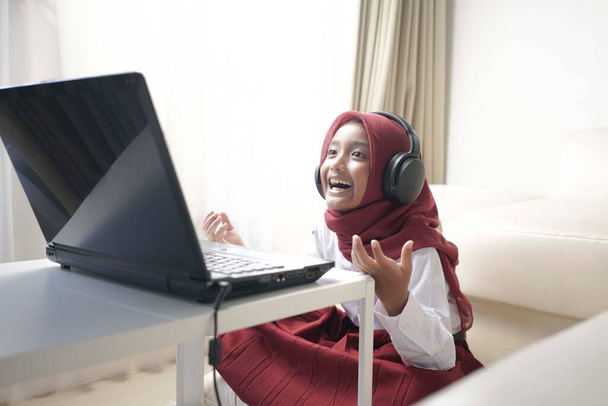 cheerful little girl wearing red hijab doing something on her laptop with headphone - Foto, imagen